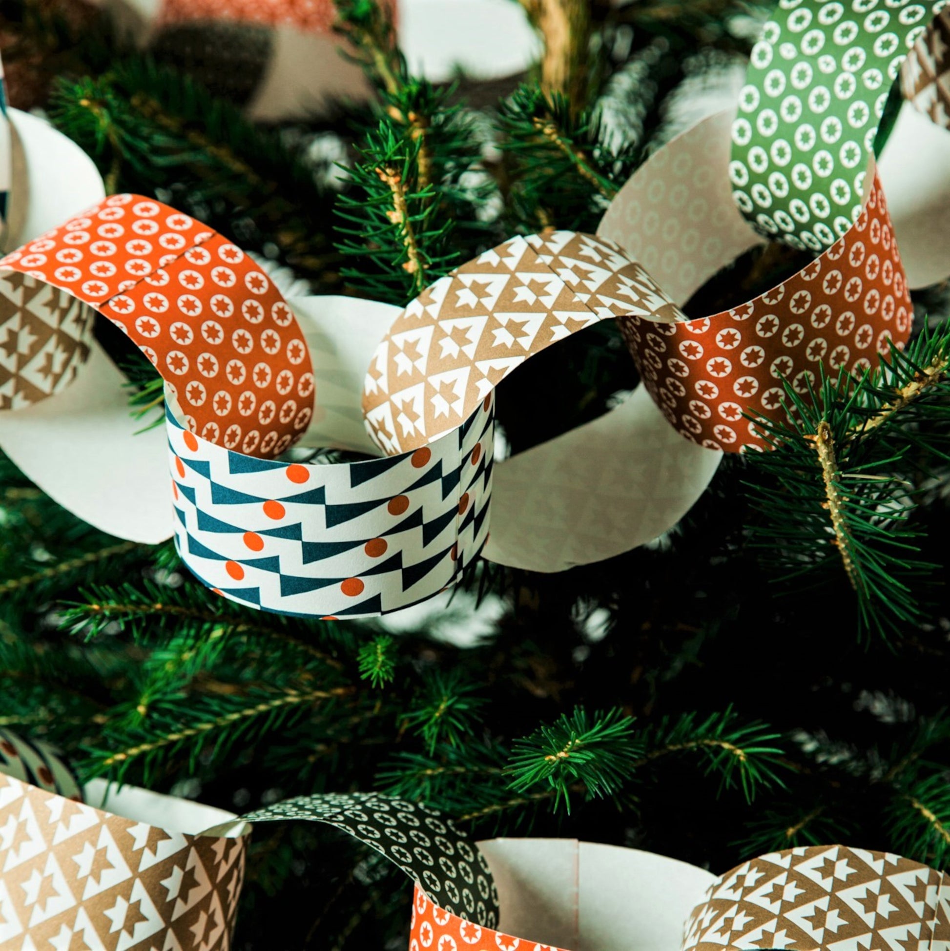 A paperchain of different patterned geometric papers in colours of gold, red, olive and blue, pictured decorating a christmas tree