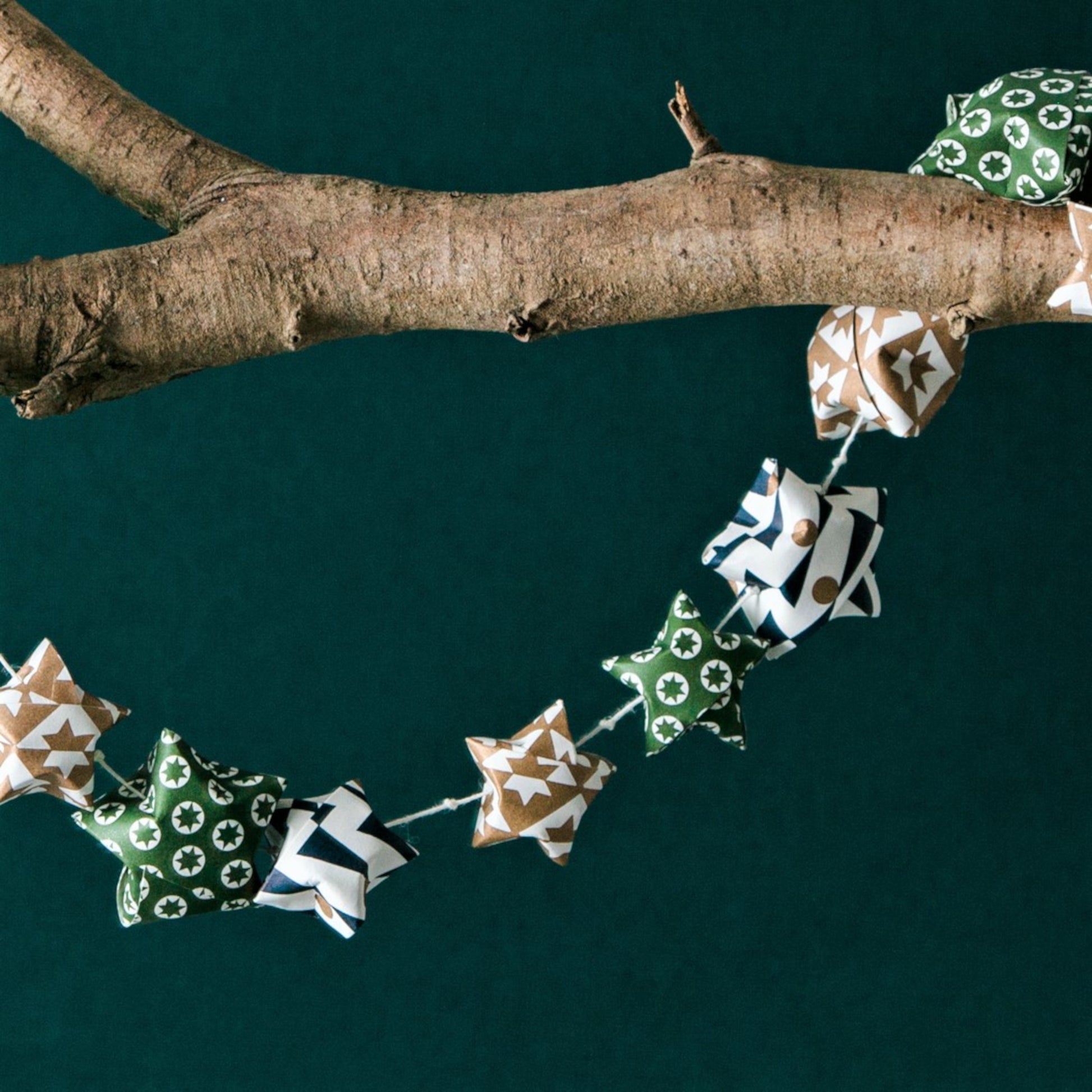 A garland of small paper origami patterned stars "lucky stars" in green, gold and dark blue, hanging from a branch
