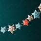 A garland of small paper origami patterned stars "lucky stars" in red, gold and aqua, pictured hanging against a dark backdrop
